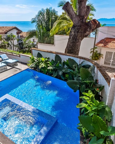 Beachfront resort pool with blue loungers, tropical plants, ocean view, and white colonial-style buildings