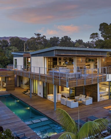 Modern two-story house with pool and wooden deck overlooking forested hills at dusk