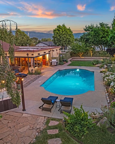 Backyard with illuminated swimming pool, lounge chairs, patio, and house with mountains visible at dusk