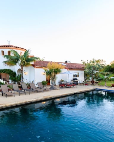 Spanish-style white house with terracotta roof and circular tower overlooking rectangular swimming pool, lounge chairs, and palm-lined grounds