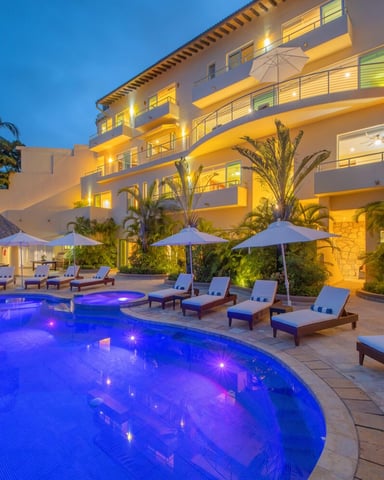 Illuminated resort pool at dusk with lounge chairs, umbrellas, and multi-story hotel building surrounded by palm trees