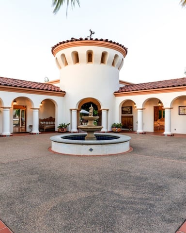 Spanish-style villa with white stucco walls, central tower, arched colonnade, courtyard fountain, and palm trees.