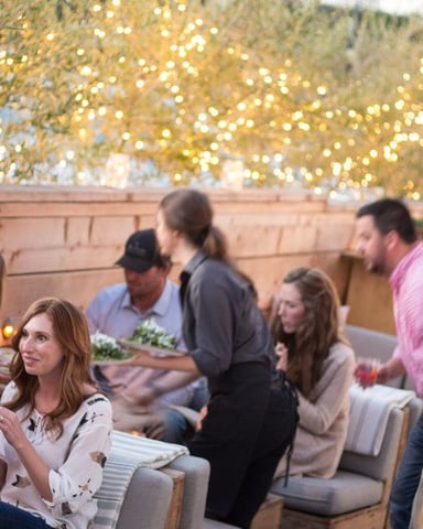 Group of people gathered in covered outdoor seating area decorated with golden garland and string lights