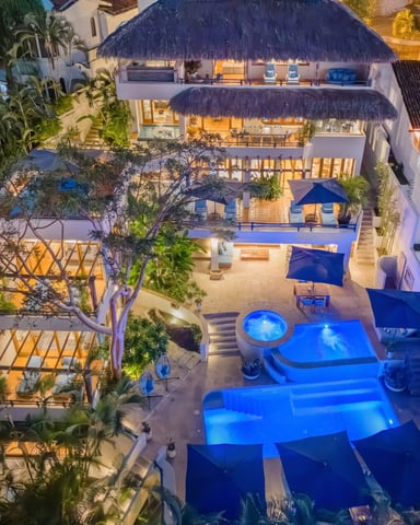Aerial view of luxury resort with illuminated pools, thatched-roof buildings, and courtyard at dusk