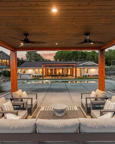 Covered patio with seating area, firepit, and view of illuminated modern house with pool at dusk