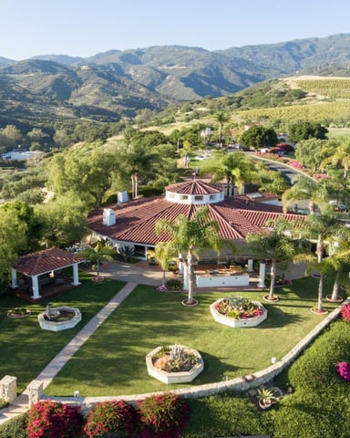Aerial view of Spanish-style estate with red-tile roofs, manicured gardens, and magenta bougainvillea overlooking vineyard-covered hills and mountains.