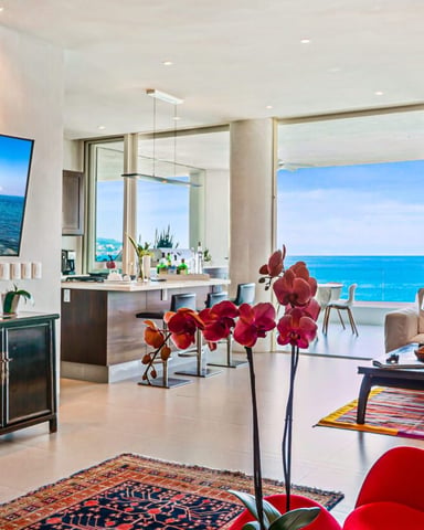 Modern open-plan living room with red chairs, ocean view through floor-to-ceiling windows, and red orchid flowers on display.