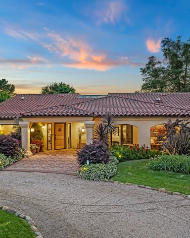 Modern two-story house with illuminated interior, wooden deck, swimming pool, and lounge chairs at dusk