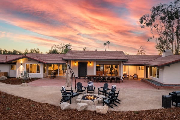 Modern single-story home with white stucco exterior, red tile roof, and lit interior at sunset. Courtyard features firepit with black Adirondack chairs, patio