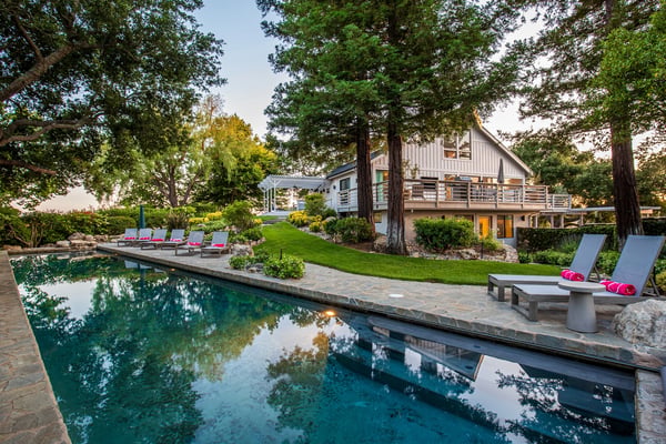 Modern white house with expansive wooden deck, lounge seating, fire pit, pool, and mature trees at dusk
