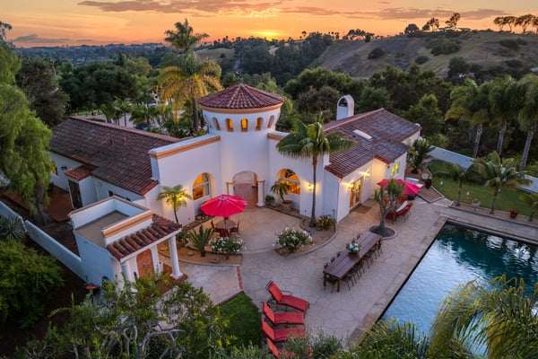 Spanish-style villa with white walls and red tile roof, pool, patio with red umbrellas, surrounded by palm trees and rolling hills at sunset.