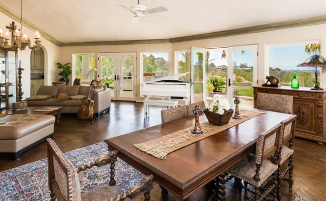 Open-plan living and dining room with wood floors, multiple glass doors opening to hillside views, ornate wooden furniture, and ceiling fan.