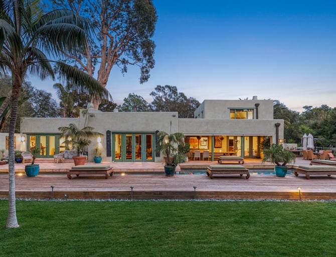 Modern concrete house with illuminated windows, wooden deck, potted palms, and manicured lawn at dusk